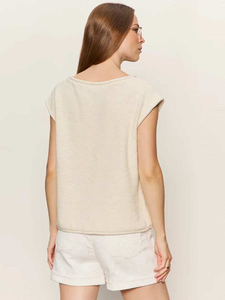 A woman with straight, light brown hair wears glasses and the Zertz EASY SWEATER TEE in cream, a relaxed-fit sleeveless knit top, paired with patterned white shorts. She faces away from the camera against a plain light background.