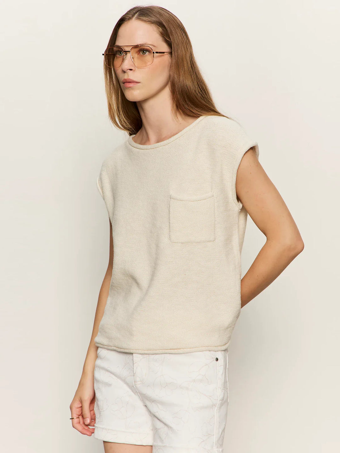 A person with long light brown hair and glasses wears the Zertz EASY SWEATER TEE, a relaxed fit sleeveless beige knit top with chest pocket, paired with light shorts, standing against an off-white background.