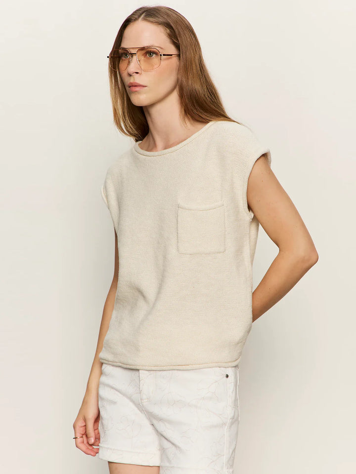 A person with long light brown hair and glasses wears the Zertz EASY SWEATER TEE, a relaxed fit sleeveless beige knit top with chest pocket, paired with light shorts, standing against an off-white background.