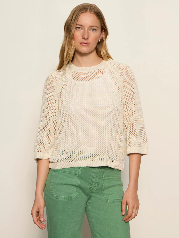 A woman with long blonde hair models the Zertz METALLIC SWEATER, a loose cream knit with three-quarter sleeves, paired with high-waisted green pants. She stands against a light background, gazing at the camera.