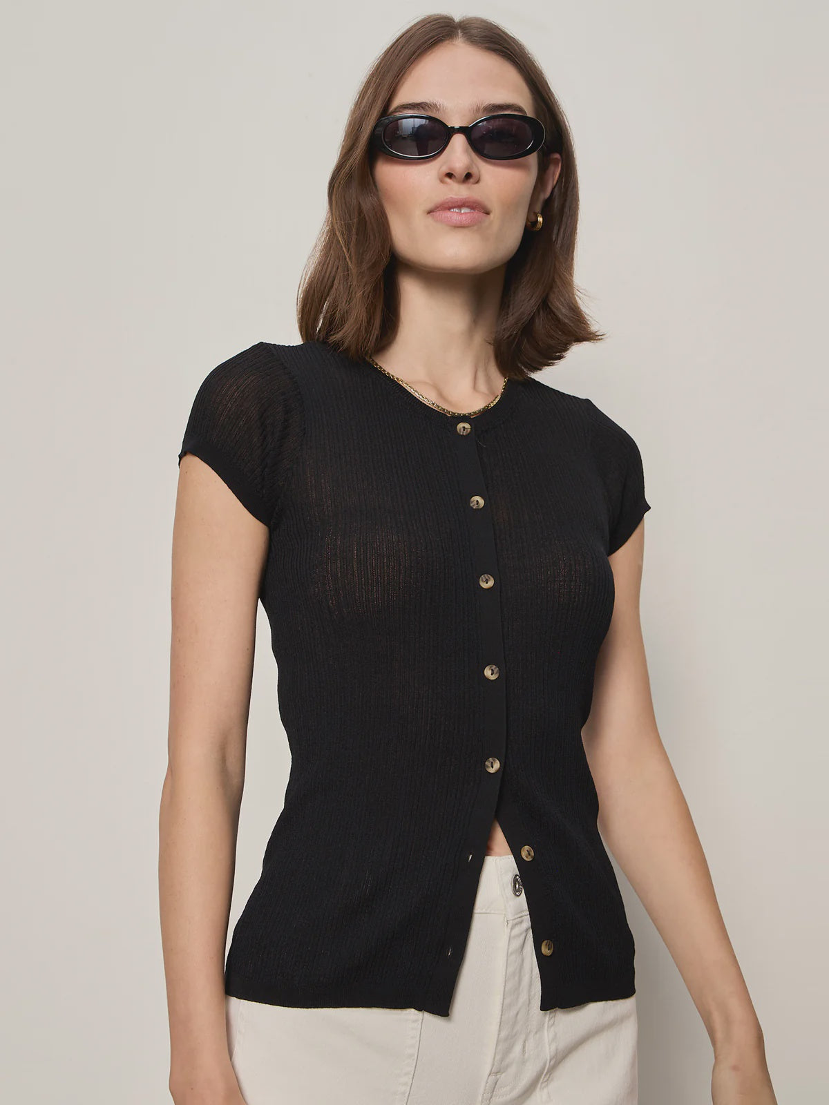 A woman stands confidently against a plain light background, wearing black sunglasses, off-white pants, and the Zertz SOFT FOCUS RIBBED TOP in black.