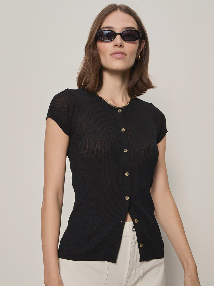 A woman stands confidently against a plain light background, wearing black sunglasses, off-white pants, and the Zertz SOFT FOCUS RIBBED TOP in black.