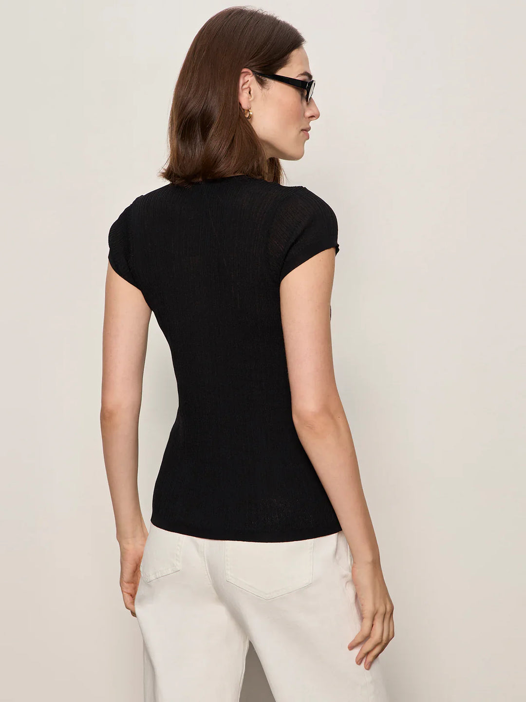 A woman with shoulder-length brown hair and sunglasses models the Zertz SOFT FOCUS RIBBED TOP, styled with high-waisted white pants, standing with her back to the camera against a plain light background.
