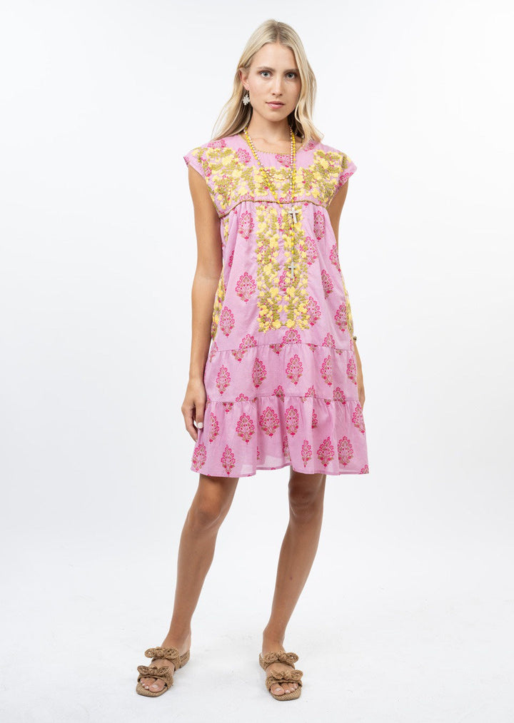 A woman with long blonde hair wears the Zertz CYNTHIA EMB FLOWER RUFFLE DRESS, a short pink floral dress featuring yellow and dark pink patterns. She accessorizes with gold earrings, a necklace, and brown sandals against a white background.