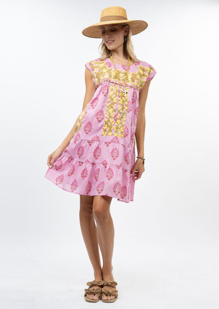 Woman stands against a plain white background wearing the Zertz CYNTHIA EMB FLOWER RUFFLE DRESS, a short-sleeved pink floral dress with yellow and pink patterns, styled with a straw hat, necklace, and brown sandals.