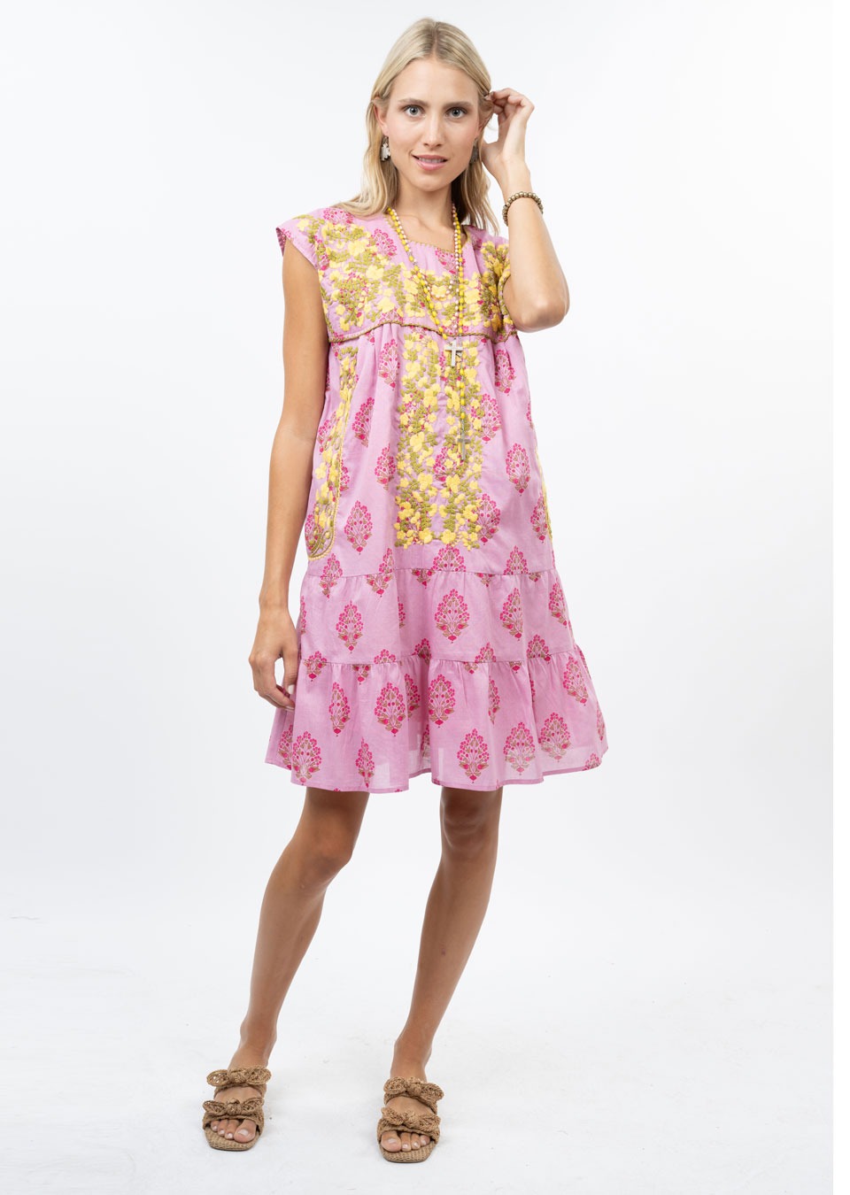 A woman with light hair stands against a white background wearing the Zertz CYNTHIA EMB FLOWER RUFFLE DRESS, a loose-fitting pink floral dress. She wears sandals and holds one hand near her face.