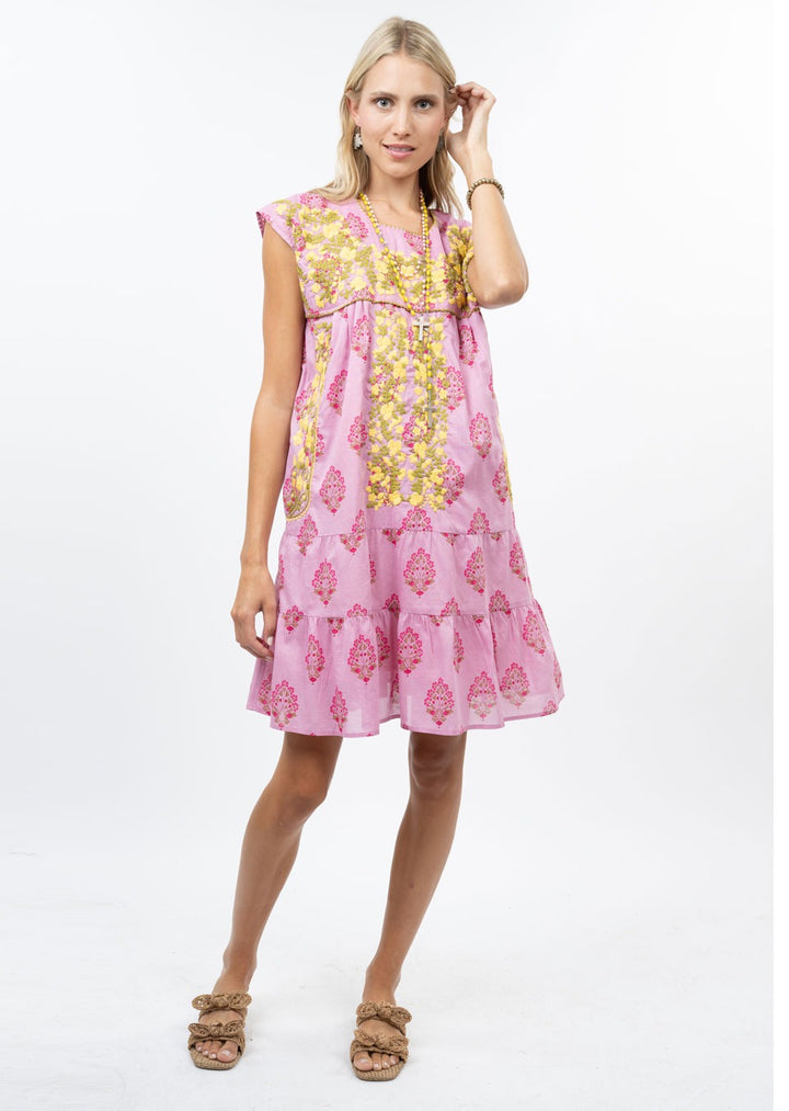 A woman with light hair stands against a white background wearing the Zertz CYNTHIA EMB FLOWER RUFFLE DRESS, a loose-fitting pink floral dress. She wears sandals and holds one hand near her face.