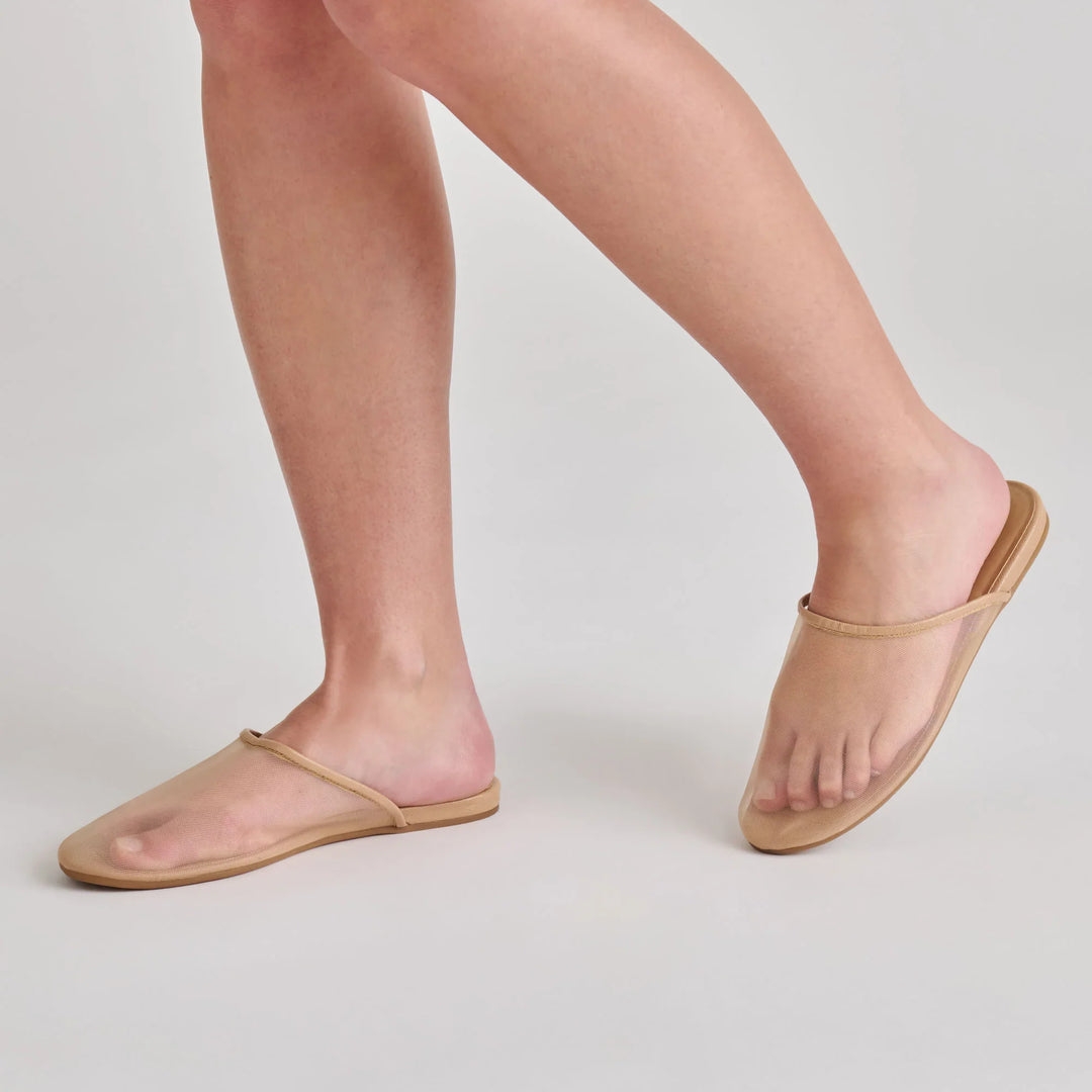 A person wears Zertz GENI beige minimalist slip-on sandals with a thin strap, exposing most of the feet. The sustainable shoes feature a simple, modern design and are shown against a light gray background.
