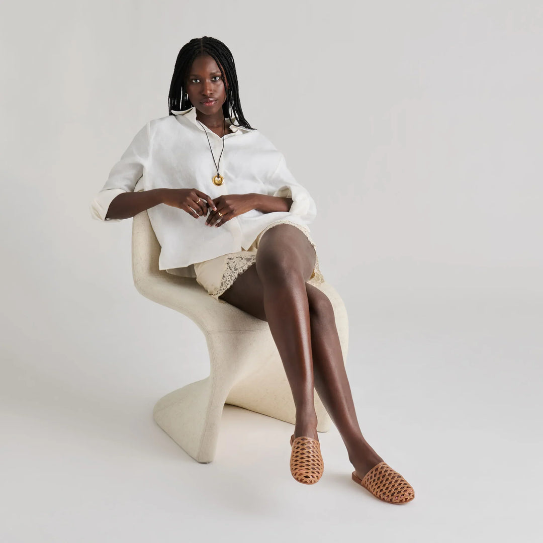 A woman with braided hair sits on a modern white chair, wearing a loose white top, light shorts, a pendant necklace, and Zertz GINNA JELLY FLATS—woven brown summer clogs made from water-based leather alternative—against a plain background.