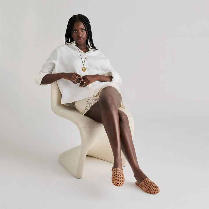 A woman with braided hair sits on a modern white chair, wearing a loose white top, light shorts, a pendant necklace, and Zertz GINNA JELLY FLATS—woven brown summer clogs made from water-based leather alternative—against a plain background.