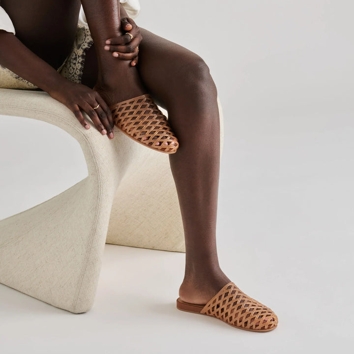 Seated on a modern cream chair, a person wears Zertz GINNA JELLY FLATS—tan woven slip-ons—paired with a light lace-trim outfit. They cross one leg and rest hands on their ankle against a plain neutral background.