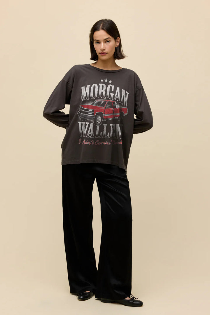 A woman poses against a beige background wearing Zertz’s MORGAN WALLEN AINT COMNG BACK MERCH LG SLV, featuring a dark graphic sweatshirt with a red truck and "Morgan Wallen," paired with black wide-leg pants and black shoes.