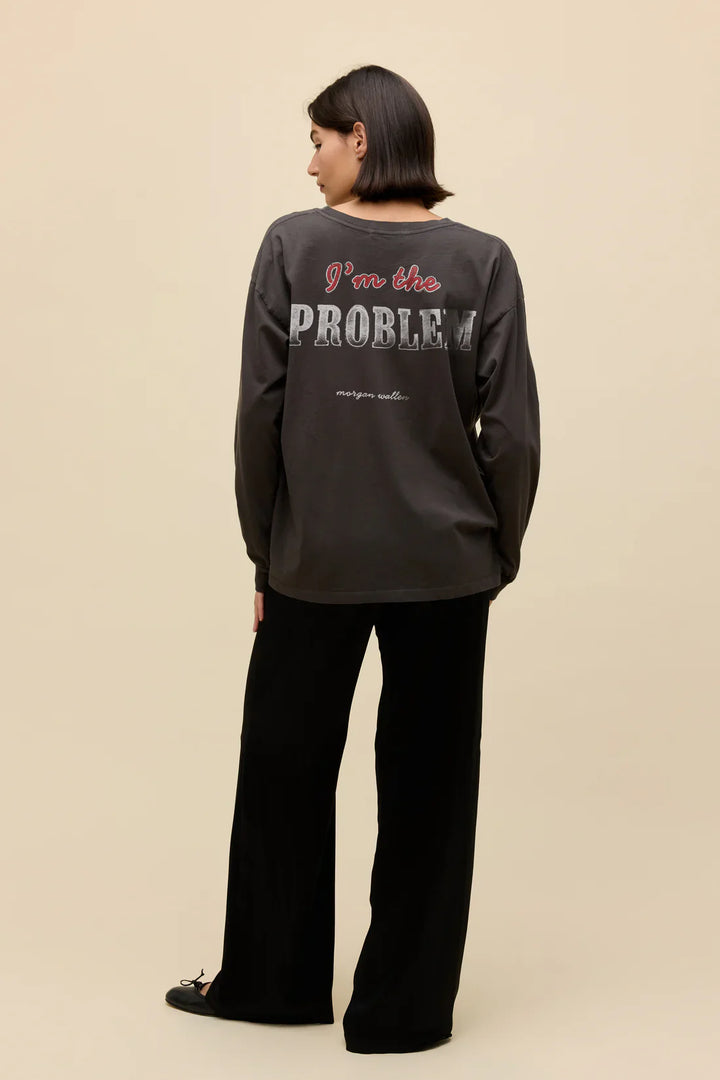 A person with short dark hair faces away, wearing the Zertz MORGAN WALLEN AINT COMNG BACK MERCH LG SLV shirt and loose black pants against a plain beige background.