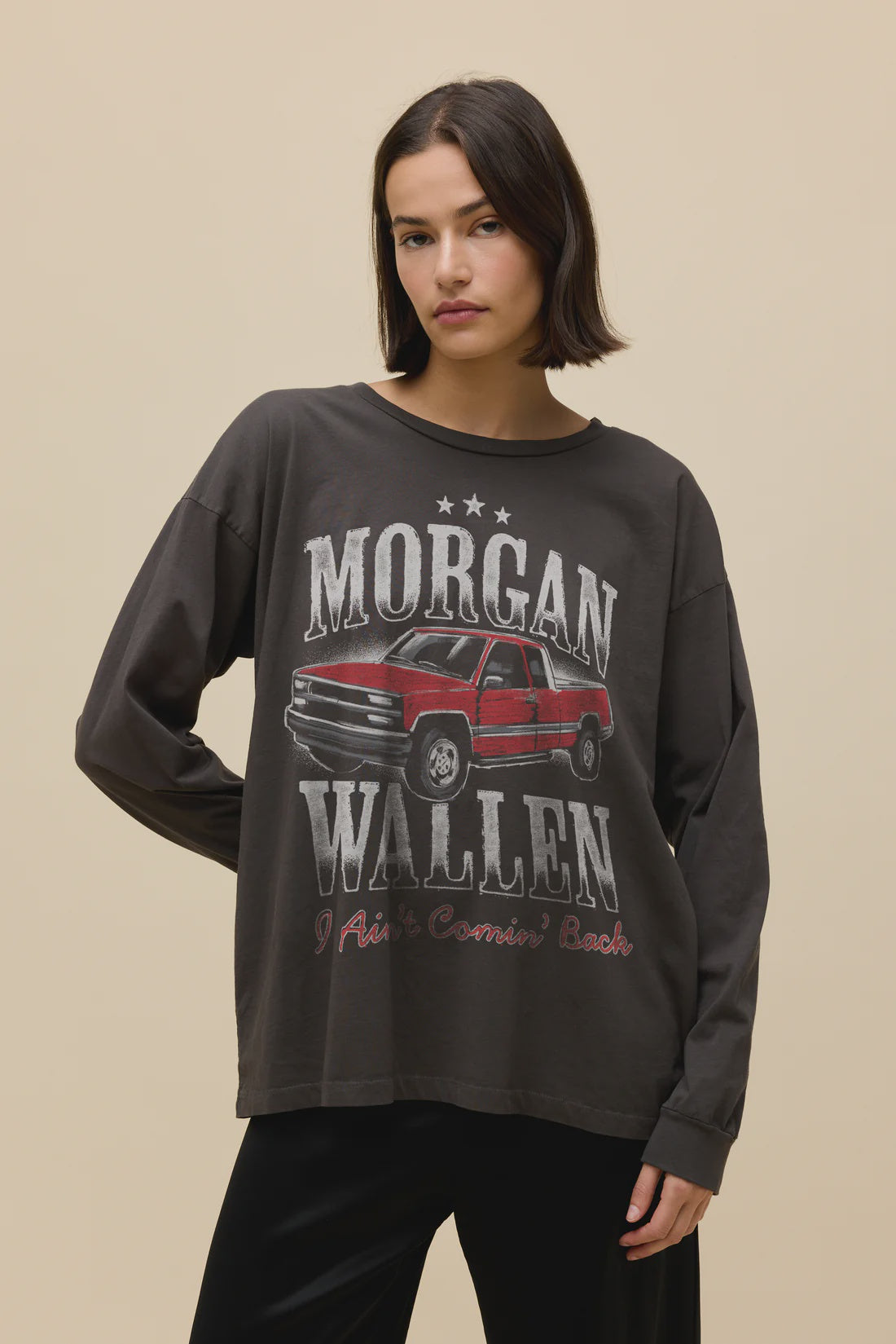 A woman with straight brown hair wears a loose, dark Zertz MORGAN WALLEN AINT COMNG BACK MERCH LG SLV shirt featuring a red pickup truck and text, standing against a plain beige background.