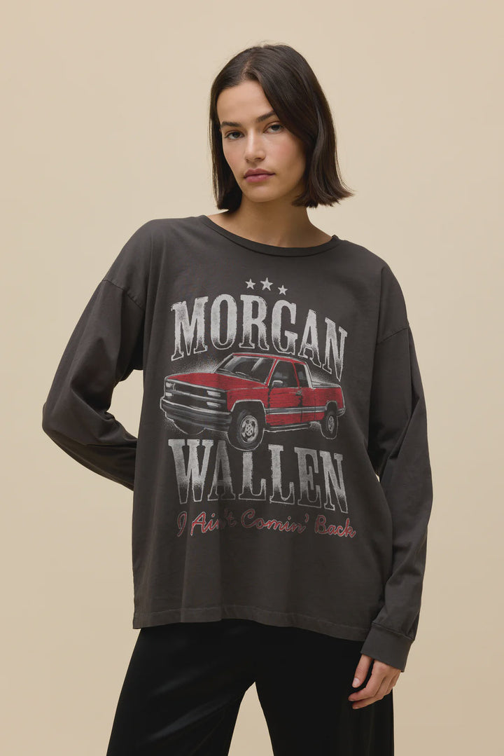 A woman with straight brown hair wears a loose, dark Zertz MORGAN WALLEN AINT COMNG BACK MERCH LG SLV shirt featuring a red pickup truck and text, standing against a plain beige background.