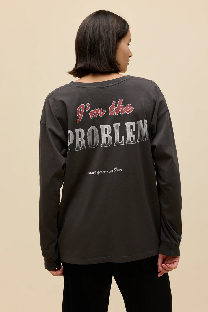 A woman with short brown hair stands with her back to the camera, wearing a Zertz MORGAN WALLEN AINT COMNG BACK MERCH LG SLV— a dark long-sleeve shirt—against a plain beige background.