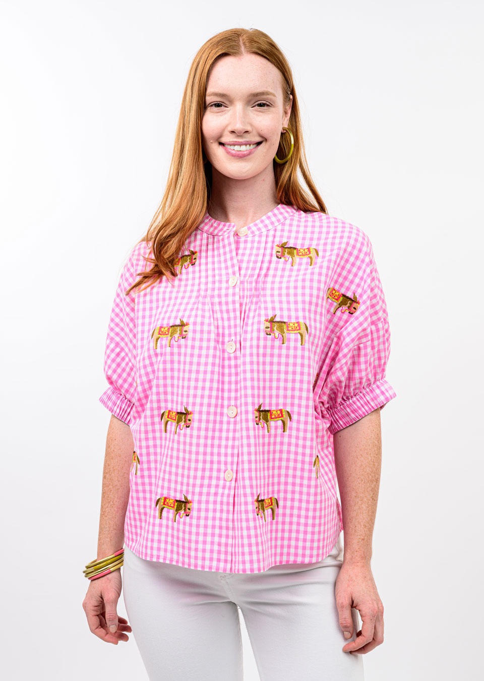 A woman with long red hair smiles at the camera, wearing the Zertz DONKEY EMB TOP—a pink and white checkered shirt with embroidered cows—styled with white pants, gold hoop earrings, and bracelets against a plain white background.