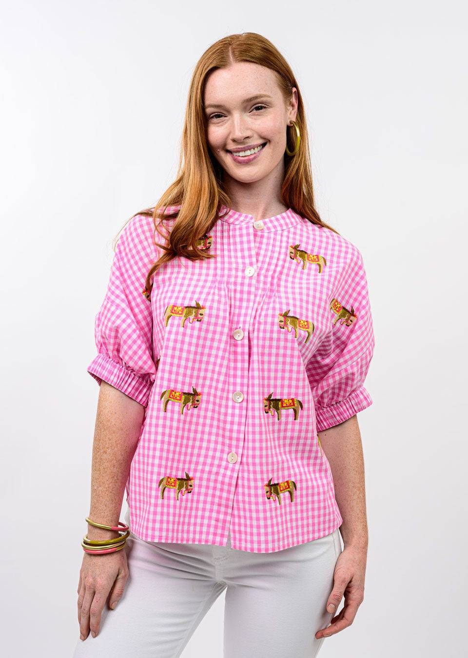 A smiling woman with long red hair wears the Zertz DONKEY EMB TOP—a whimsical pink and white checkered blouse with embroidered tigers—paired with white pants and gold hoop earrings, standing against a plain white background.