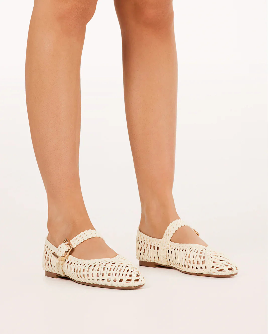 A person stands on a white background wearing Zertz FRANKY FLAT cream-colored raffia flats with ankle straps, rounded toes, and a chic woven design.