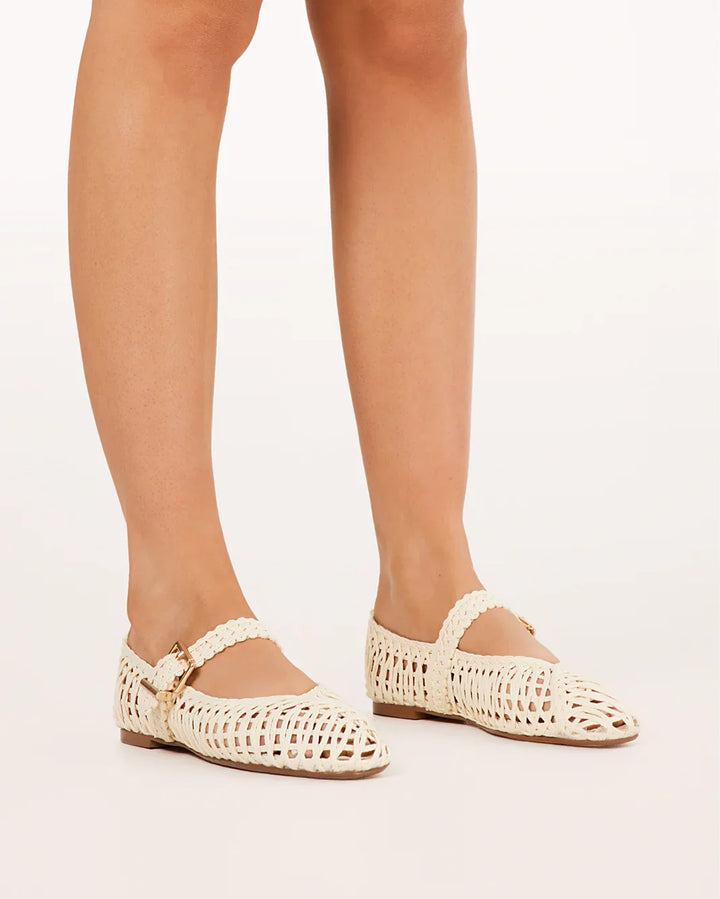 A person stands on a white background wearing Zertz FRANKY FLAT cream-colored raffia flats with ankle straps, rounded toes, and a chic woven design.