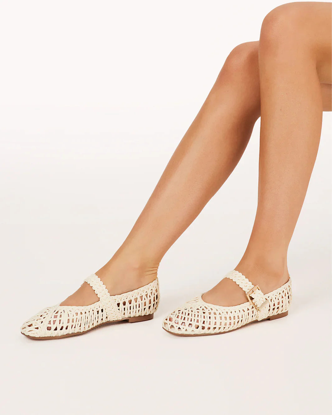 A person with crossed legs wears the FRANKY FLAT by Zertz, featuring a chic woven raffia design and buckled strap, shown against a plain white background.