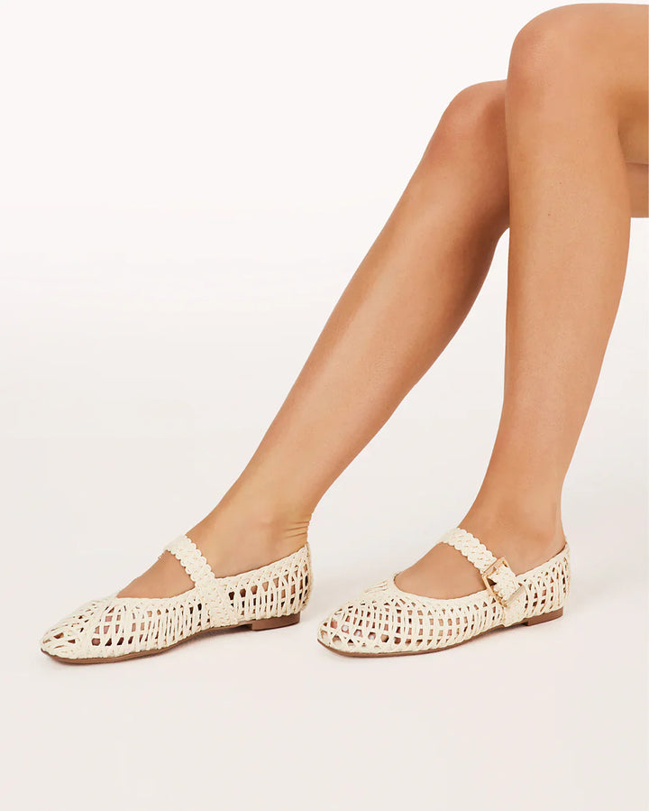 A person with crossed legs wears the FRANKY FLAT by Zertz, featuring a chic woven raffia design and buckled strap, shown against a plain white background.