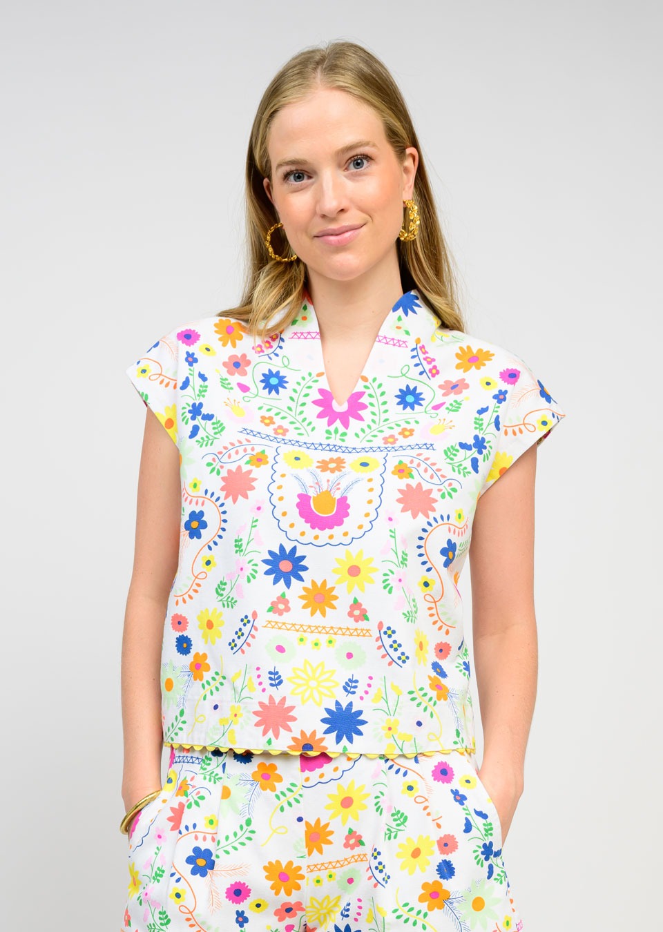 A woman with long blonde hair and gold hoop earrings wears the Zertz FLORES TOP, a floral design crafted from breathable fabric for an effortlessly feminine look against a plain light background.