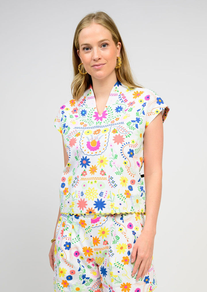 A woman with long blonde hair wears the Zertz FLORES TOP, a floral, breathable piece with vibrant flowers and birds. She stands against a light gray background, smiling slightly at the camera for a feminine look.