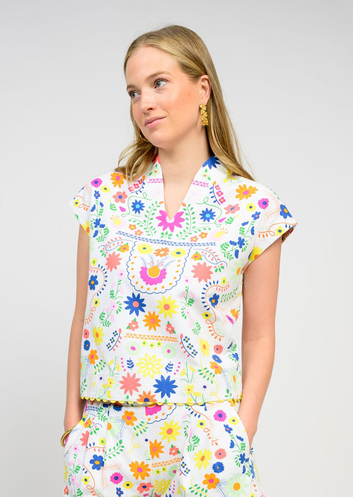 A woman with long blonde hair wears the Zertz FLORES TOP, a white short-sleeve floral piece paired with matching colorful pants. She looks slightly to the side with her hands in her pockets, embodying a feminine style.