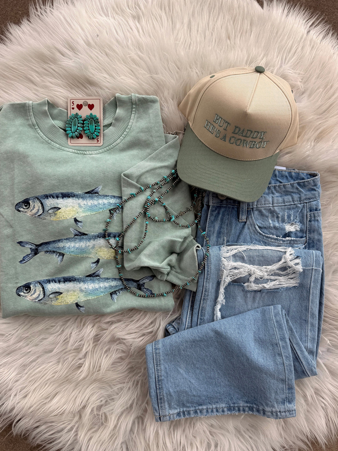 A flat lay features the Loca on Main Three Sardines Sweatshirt, light blue ripped jeans, a beige-green "But Daddy, He’s a Cowboy" cap, turquoise earrings, and beaded necklaces for a vintage vibe on a fuzzy white surface.