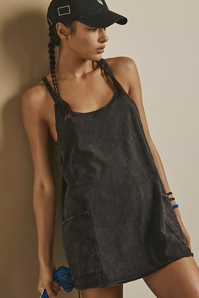 A woman with two braids, wearing a black cap and the Zertz HOT SHOT MINI DRESS—a loose, sleeveless dark gray athleisure dress with a large pocket—stands against a light wall, holding a blue textured object.