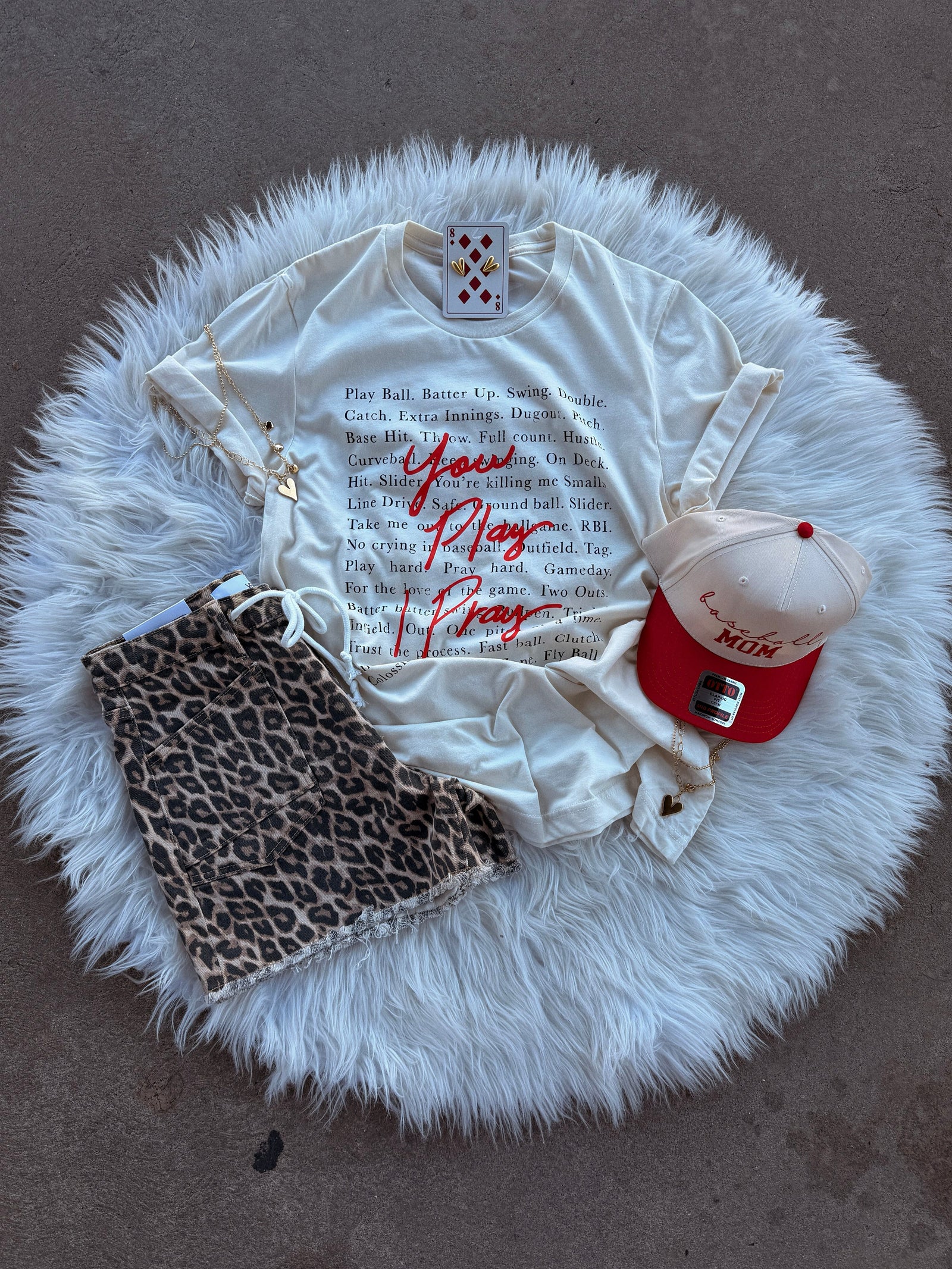 A flat lay features the Loca on Main "You Play, I Pray" tee, leopard print shorts, a cream and red The MUK baseball cap, a card, and accessories arranged on a white fur rug over a brown floor.