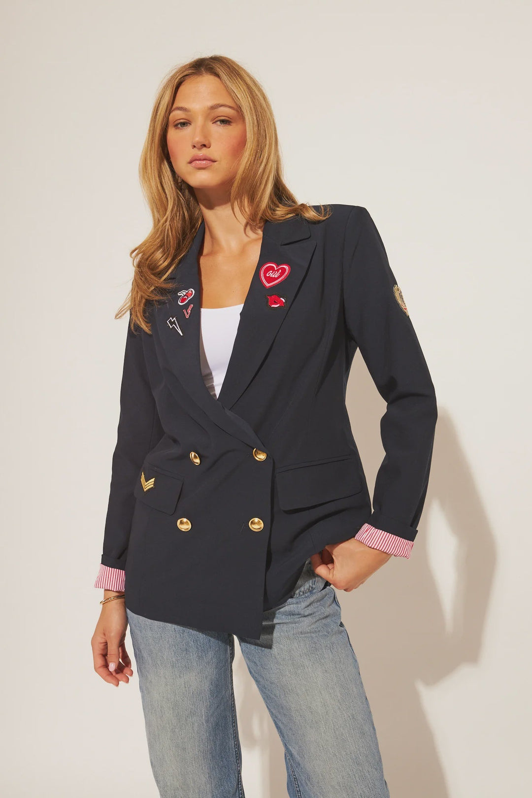 A woman with light brown hair models the Zertz BLAINE BLAZER, a navy blazer with gold buttons, colorful heart patches, and pink striped cuffs, layered over a white top and blue jeans against a plain light background.