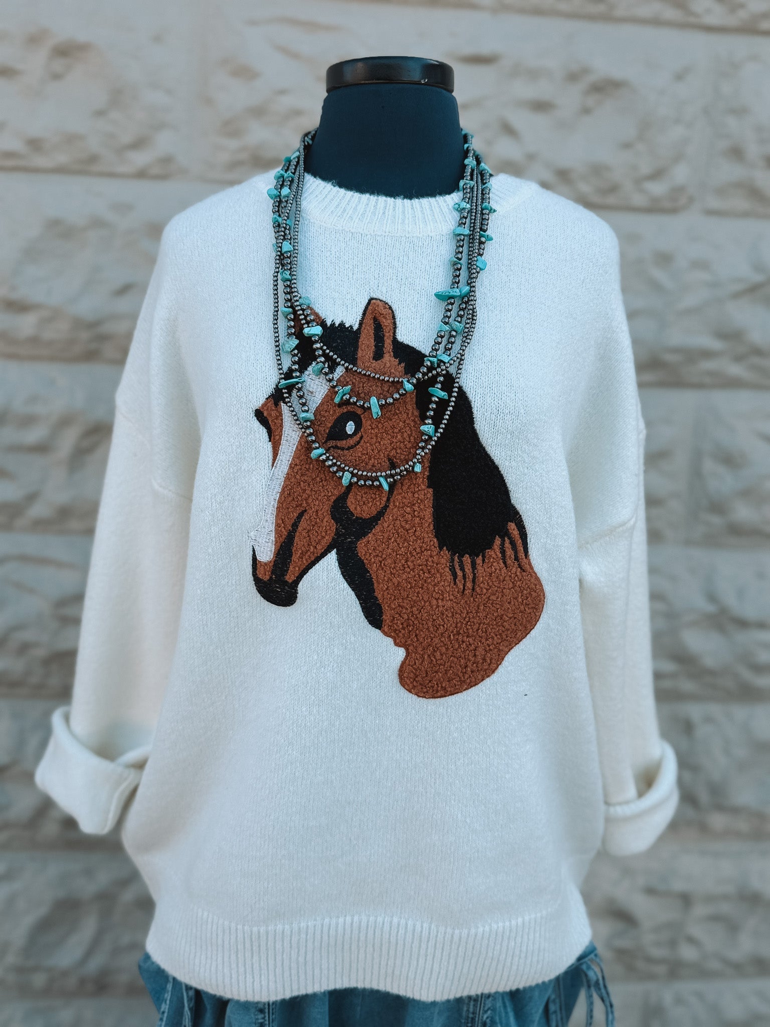 A mannequin models the Loca on Main Pony Sweater, an ivory knit with a bold brown and black horse head motif. Turquoise and beaded necklaces add flair, all set against a textured stone wall backdrop.