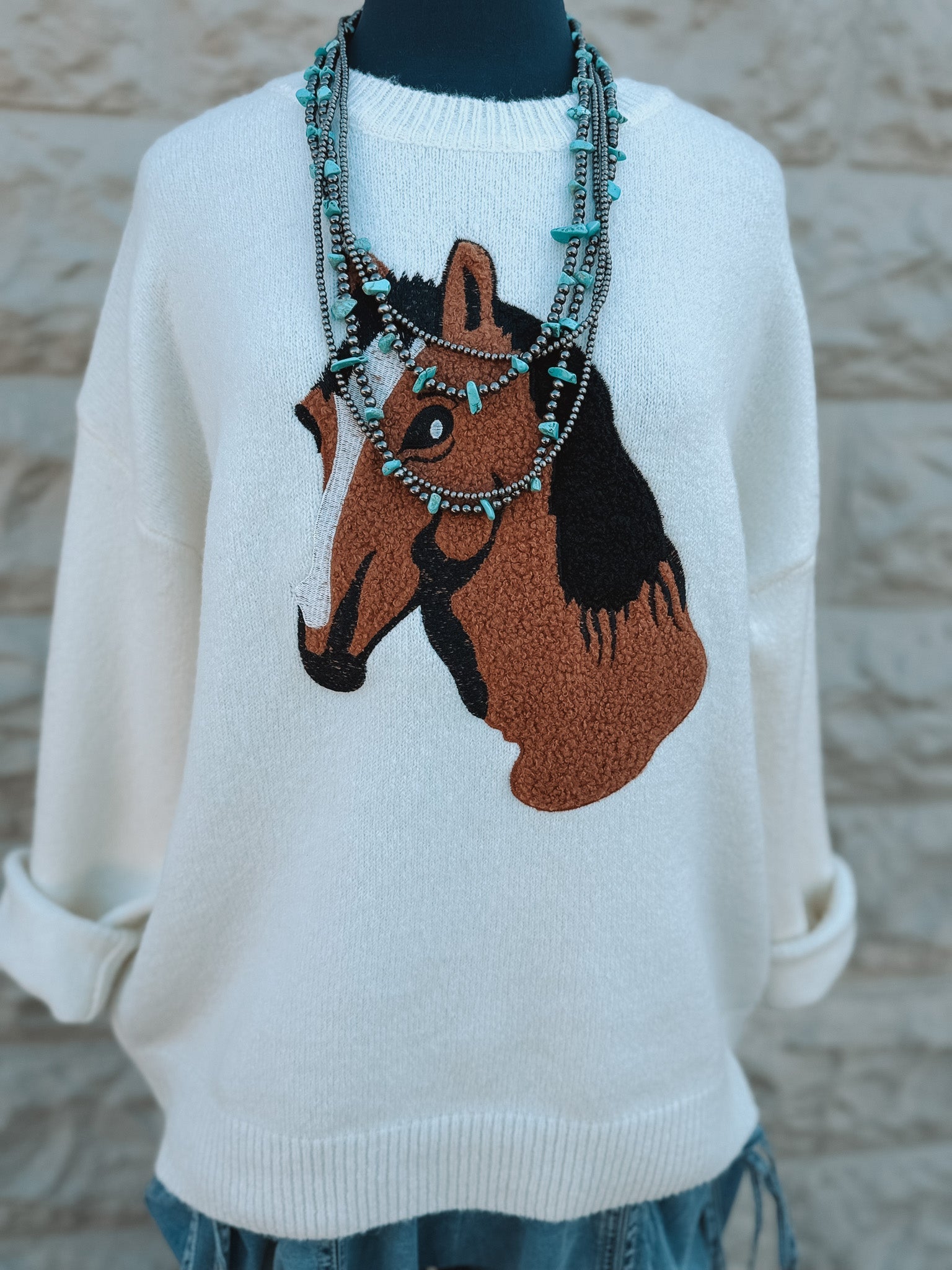 The Pony Sweater by Loca on Main features an ivory knit with a striking brown and black horse head design, shown on a mannequin styled with layered turquoise beaded necklaces.