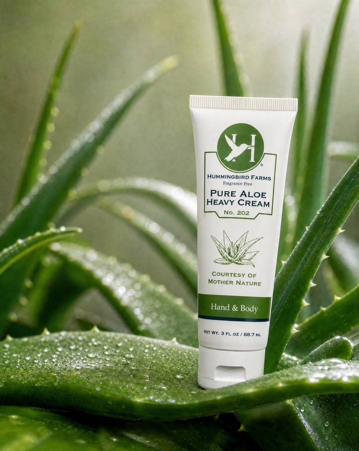 A 3oz tube of Hummingbird Farms Pure Aloe Heavy Cream No. 202, a gentle fragrance-free moisturizer, stands upright among dewy aloe vera leaves against a lush green background.