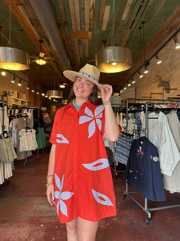 A woman smiles inside a clothing store, holding her straw hat and wearing the Zertz SHORT SLEEVE FLORAL EMBROIDERED DRESS in red with large blue and white flowers. Clothing racks and hanging lights surround her.
