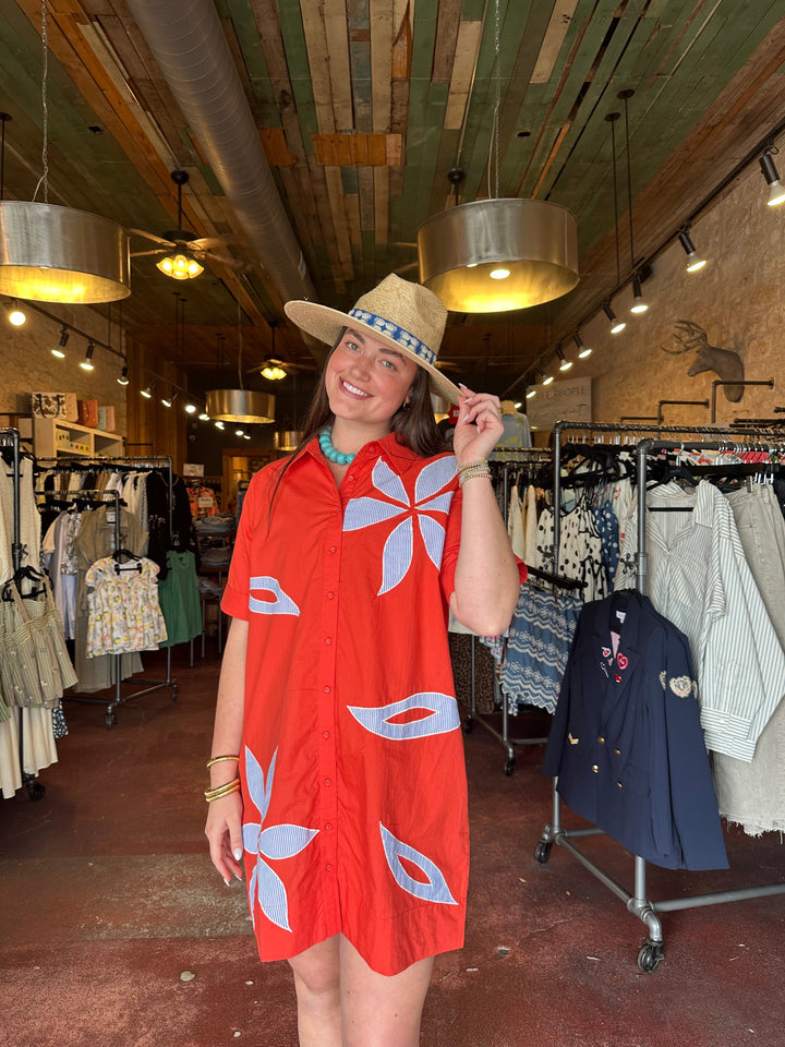 A smiling woman wearing the Zertz SHORT SLEEVE FLORAL EMBROIDERED DRESS and a straw hat stands in a boutique clothing store, surrounded by racks of clothes and warm decorative lighting overhead.