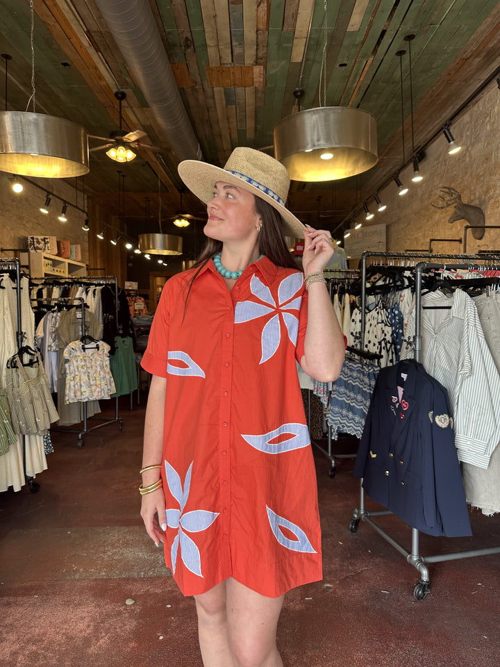 A woman wearing the Zertz SHORT SLEEVE FLORAL EMBROIDERED DRESS and a straw hat stands in a boutique with clothing racks and hanging lights, glancing to her left amid the rustic shop interior.