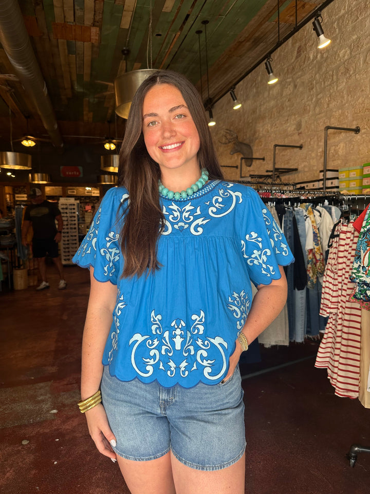 A woman with long brown hair smiles in a clothing store, wearing the Zertz EMROIDERED DETAILED TOP and denim shorts—perfect inspiration for a versatile wardrobe.
