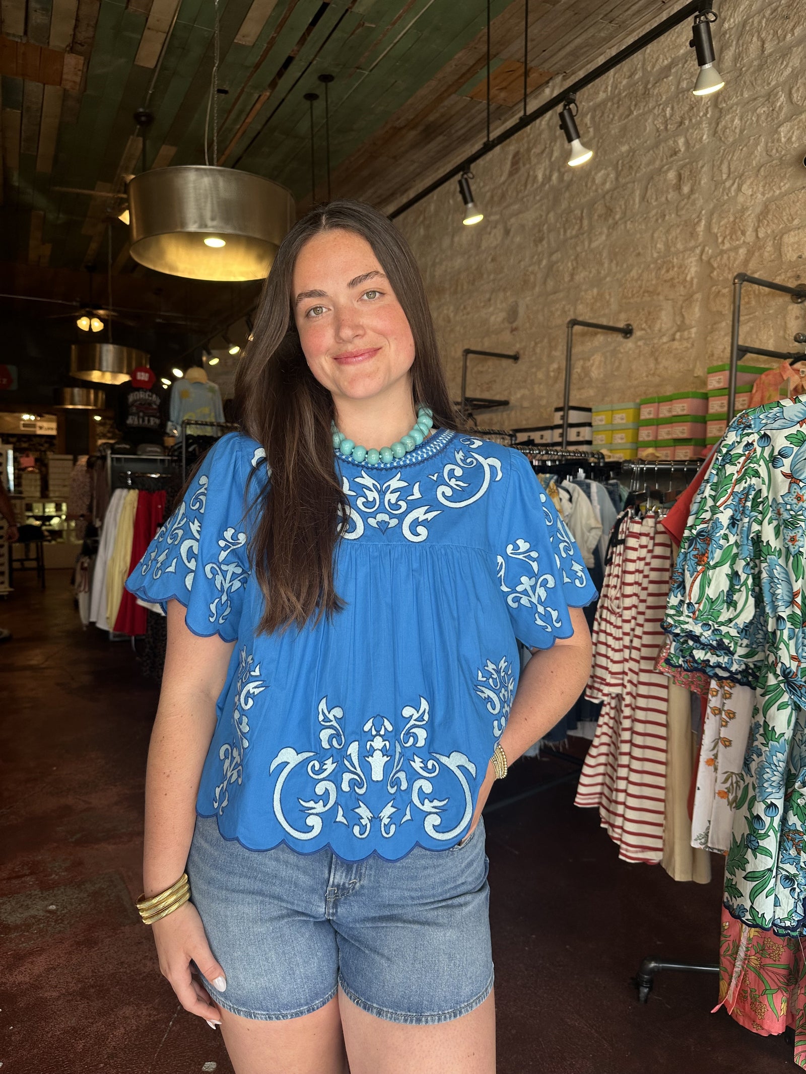 A woman with long brown hair smiles in a clothing store, wearing the Zertz EMBROIDERED DETAILED TOP and denim shorts—an ideal scene for discovering versatile styles.