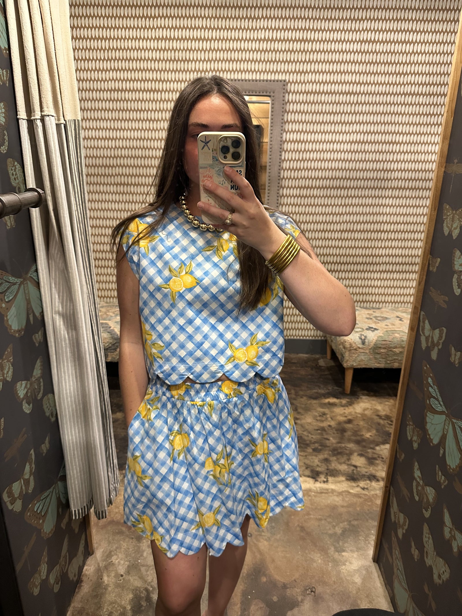 A person takes a mirror selfie in the Zertz 2 PIECE LEMON SET, featuring blue and white checks with yellow prints. The summery look is complemented by butterfly-patterned wallpaper and a matching bench in the background.