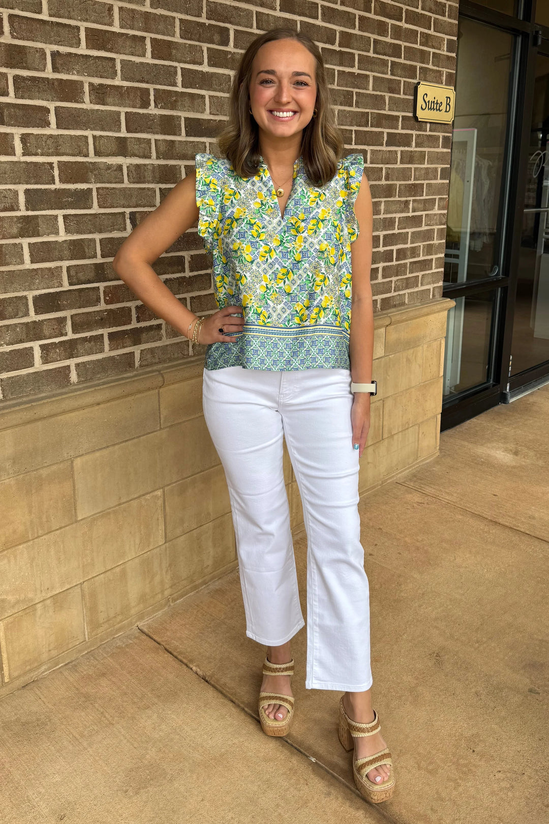 A woman with light brown hair smiles outdoors by a brick wall, wearing the Zertz PUJA TOP—a feminine lemon print blouse with ruffle sleeves—paired with white pants, tan platform sandals, a watch, and a bracelet as she rests her hand on her hip.