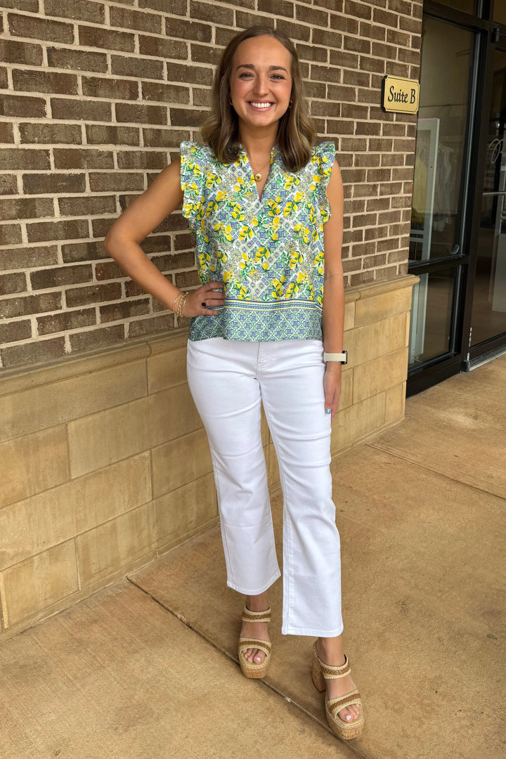 A woman with light brown hair smiles outdoors by a brick wall, wearing the Zertz PUJA TOP—a feminine lemon print blouse with ruffle sleeves—paired with white pants, tan platform sandals, a watch, and a bracelet as she rests her hand on her hip.