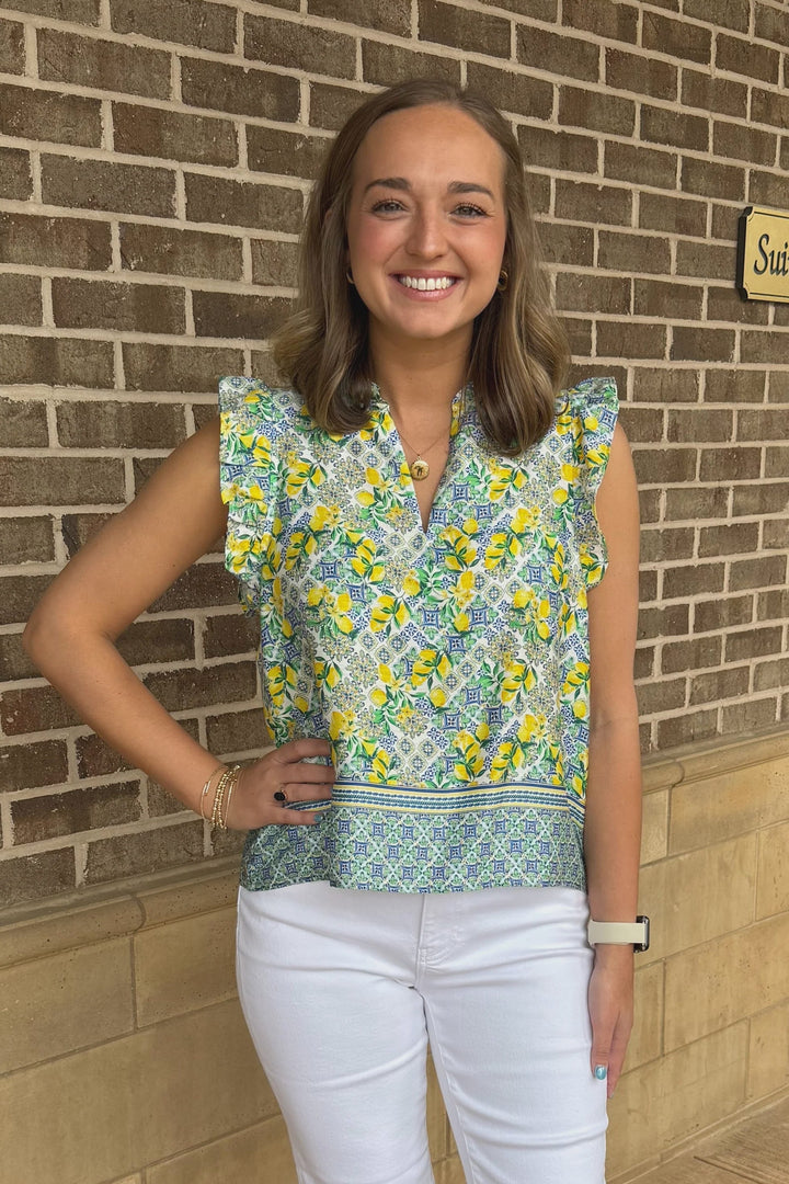 A smiling woman with shoulder-length brown hair poses in front of a brick wall, wearing the Zertz PUJA TOP—a feminine lemon print blouse with ruffle sleeves—paired with white pants, bracelets, and a smartwatch.