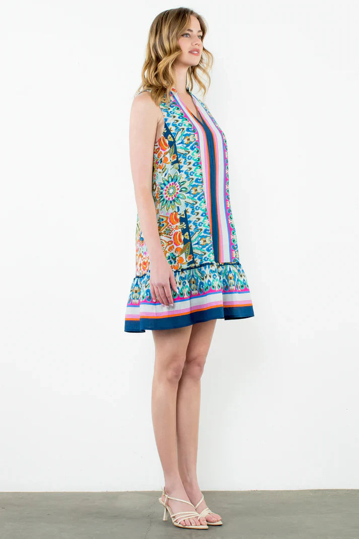 A woman stands in profile, wearing the Zertz SLVLESS V NECK PRINTED DRESS featuring a vibrant floral and geometric pattern, paired with beige strappy heels, against a plain white background.