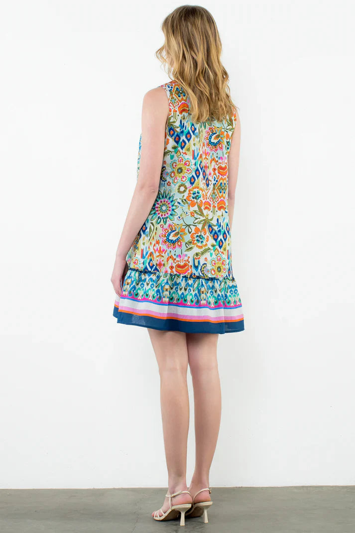 A woman with wavy blond hair models the Zertz SLVLESS V NECK PRINTED DRESS, featuring a floral print and ruffled hem, as she stands in sandals facing away against a plain white wall.