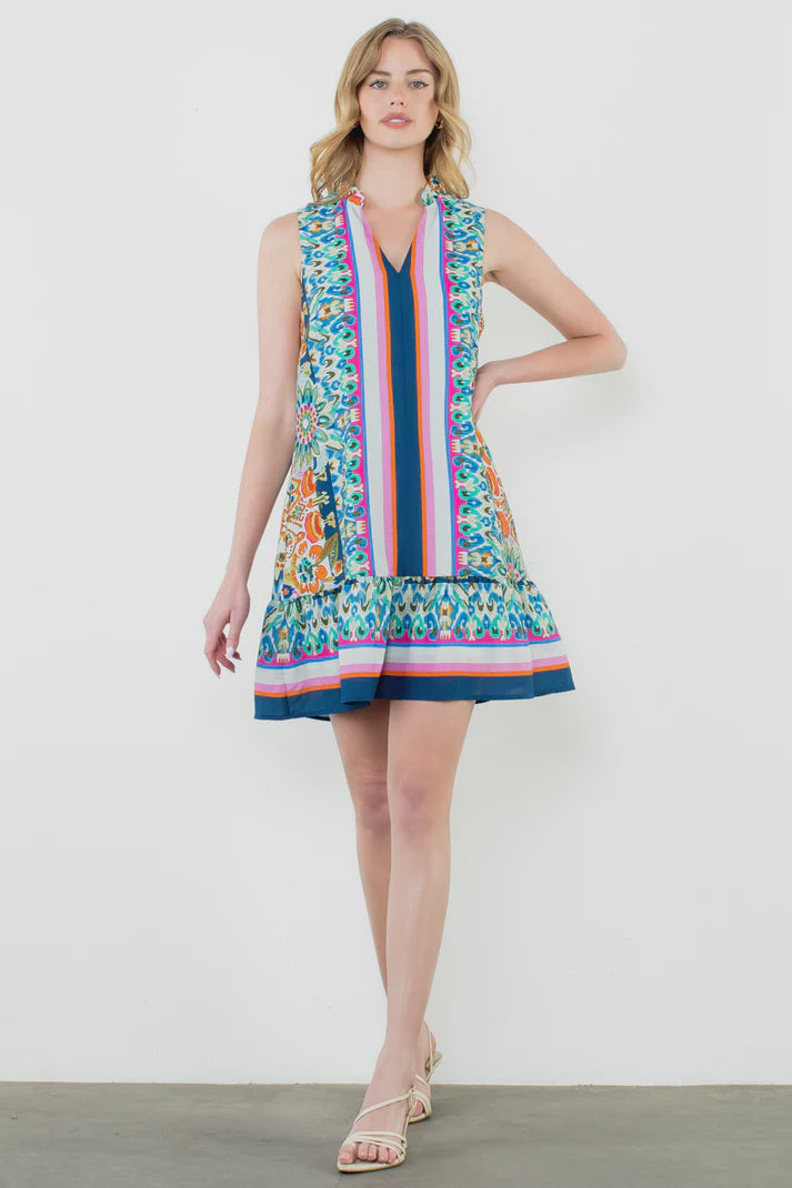 A woman with light wavy hair wears the Zertz SLVLESS V NECK PRINTED DRESS, featuring a multicolored pattern and flared hem, paired with strappy sandals. She stands against a white background with one hand on her hip.