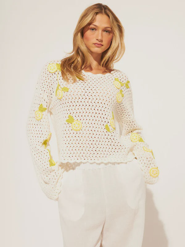 A woman with long blonde hair models the Zertz ORLANDO SWEATER TOP, a 100% cotton lightweight knit featuring yellow lemon designs, paired with white pants and set against a plain light background.