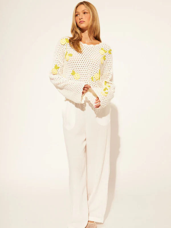 A woman with long blonde hair, looking relaxed, models the Zertz ORLANDO SWEATER TOP— a lightweight 100% cotton knit with yellow flower patterns—paired with loose white pants against a white background.